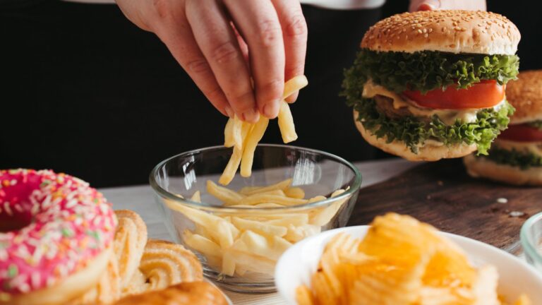 A hand reaches for a bowl of fries next to a burger, donuts, chips, and other junk food.