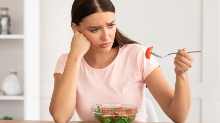 A woman frowns at the tomato on her fork from her salad. Image demonstrates how gaining weight while trying to lose it is frustrating.