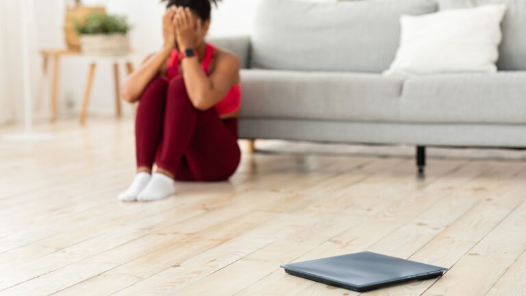 A woman sits with her head in her hands across the floor from a scale. Image demonstrates how you feel after gaining weight back.