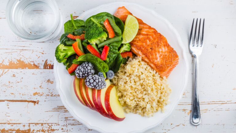 Apples, rice, salmon, vegetables, and water all sit next to each other on a table. Image demonstrates examples of a healthy diet.