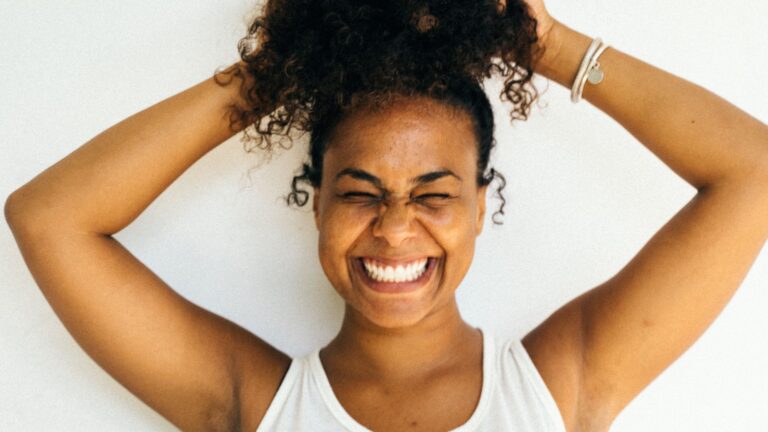 A woman is smiling widely, squeezing her eyes shut, with her arms above her head. Image demonstrates the feeling of learning how to get rid of cellulite.