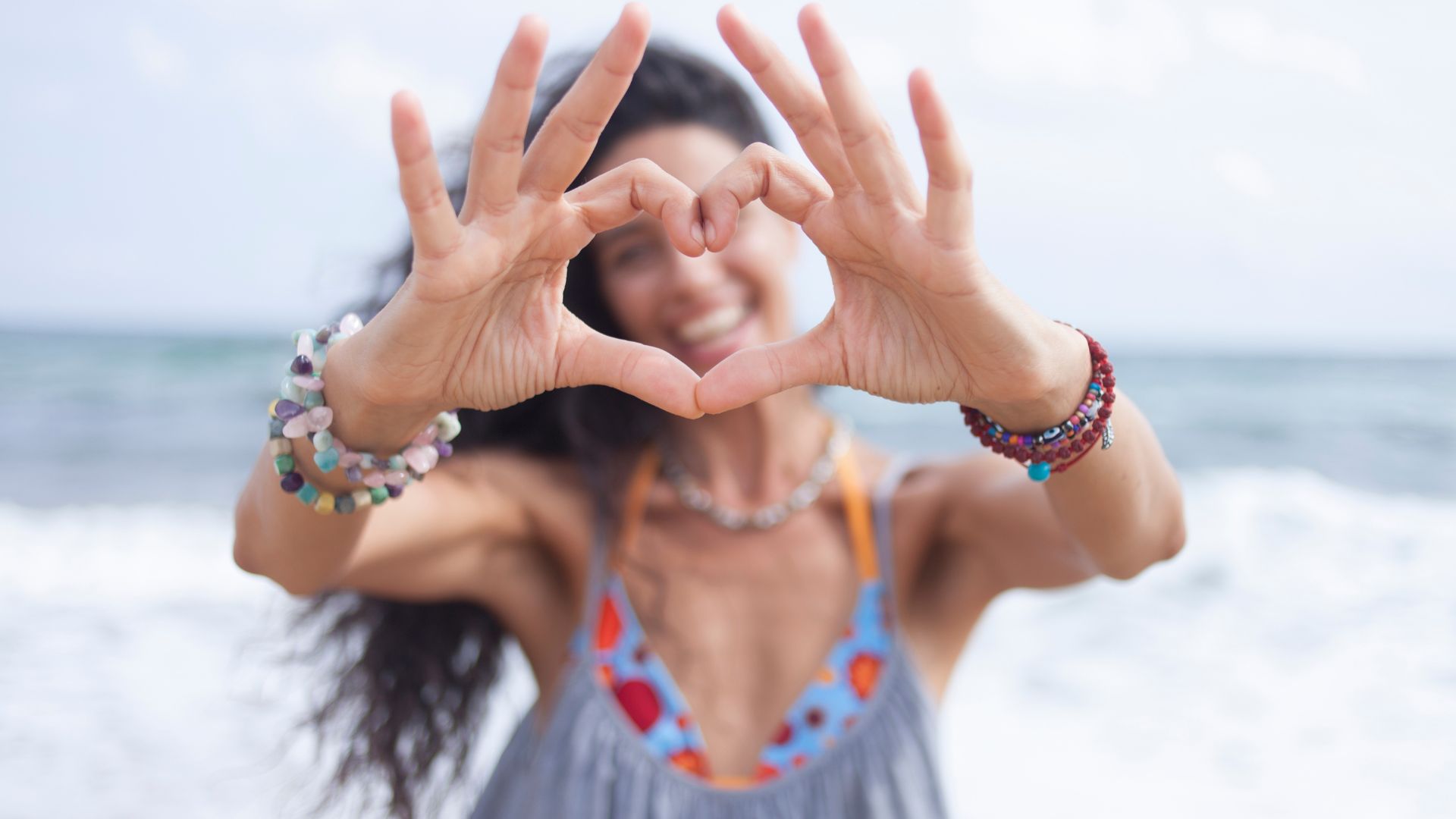 A woman smiles while making a heart with her hands, the beach in the background.