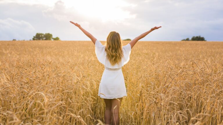 A woman stands in the middle of a corn field in a white dress, her arms raised up in the air joyfully. Image demonstrates the feeling of learning how to reduce inflammation in the body fast.