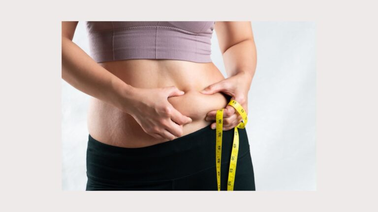 A woman holding a measuring tape pinches her love handles. Image demonstrates how hard it is to lose belly fat.