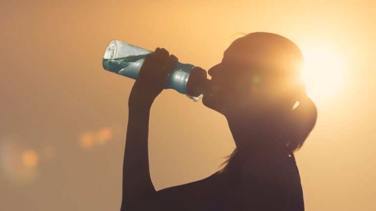 A woman drinks from a water bottle, backed by the sun. Image demonstrates a technique to lose weight quickly.