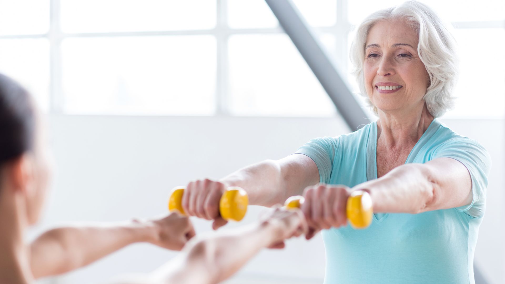 An older woman holds weights out in front of her, working out with a personal trainer. Image demonstrates how women after menopause work to lose weight.