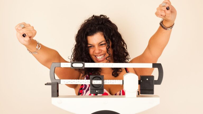 A woman smiles at the weight reading on her scale. Image demonstrates how you feel when weight loss programs work.