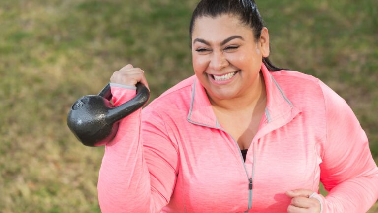 A woman smiles while lifting a kettlebell, working out outside. Image demonstrates how you feel when you keep weight off.