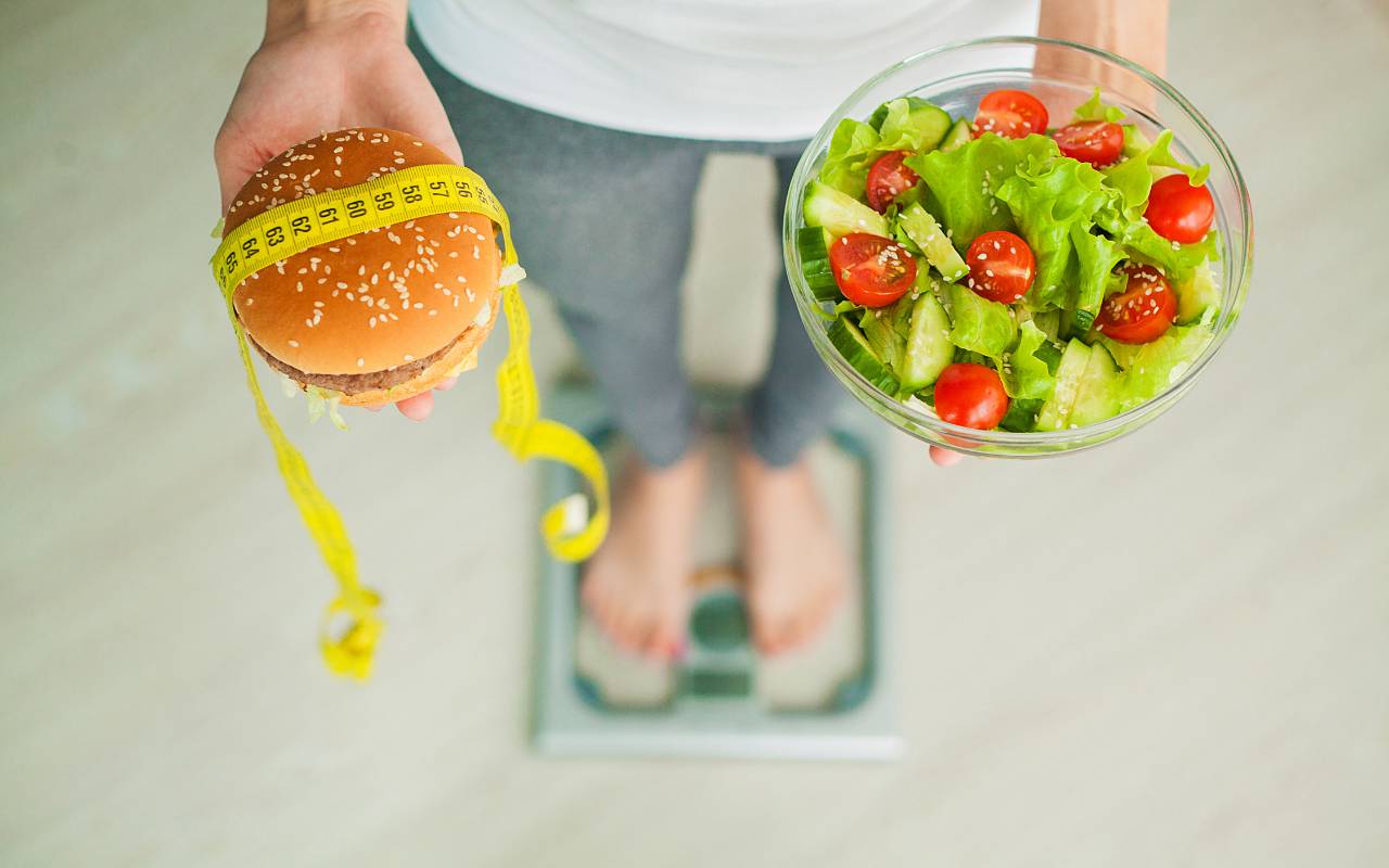 A woman standing on a scale with a burger in one hand and a salad in the other hand while she debates how many calories she should eat to lose weight.