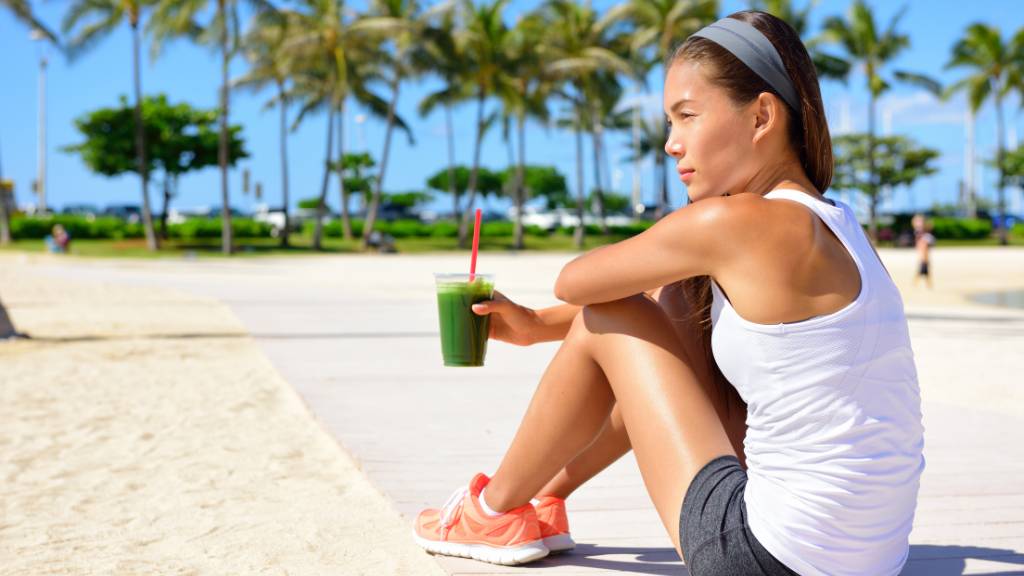 A woman sitting on a beach sidewalk, by the sand, drinking a healthy drink and debating her calorie deficit diet.