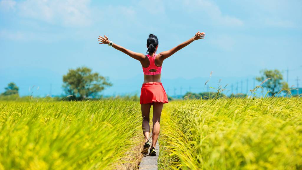 The back of a female walking a dirt path in a meadow and raising her hands to celebrate keeping the weight off sustainably.