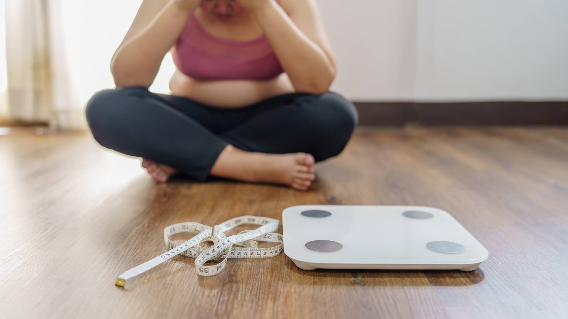 A women sits cross legged with her head in her hands. In front of her is a scale and a measuring tape. This is demonstrating why we gain weight.