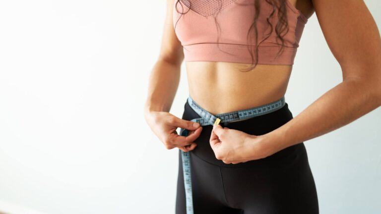 A woman stands wearing black leggings and a pink sports bra. Only her torso shows as she puts a measuring tape around her waist. She is demonstrating her goal to lose weight.