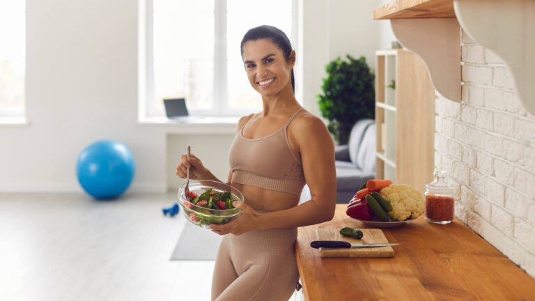 A woman wearing a tan workout outfit holds a healthy meal in her kitchen. There is a workout ball in the background. She is demonstrating a healthy way to lose weight.