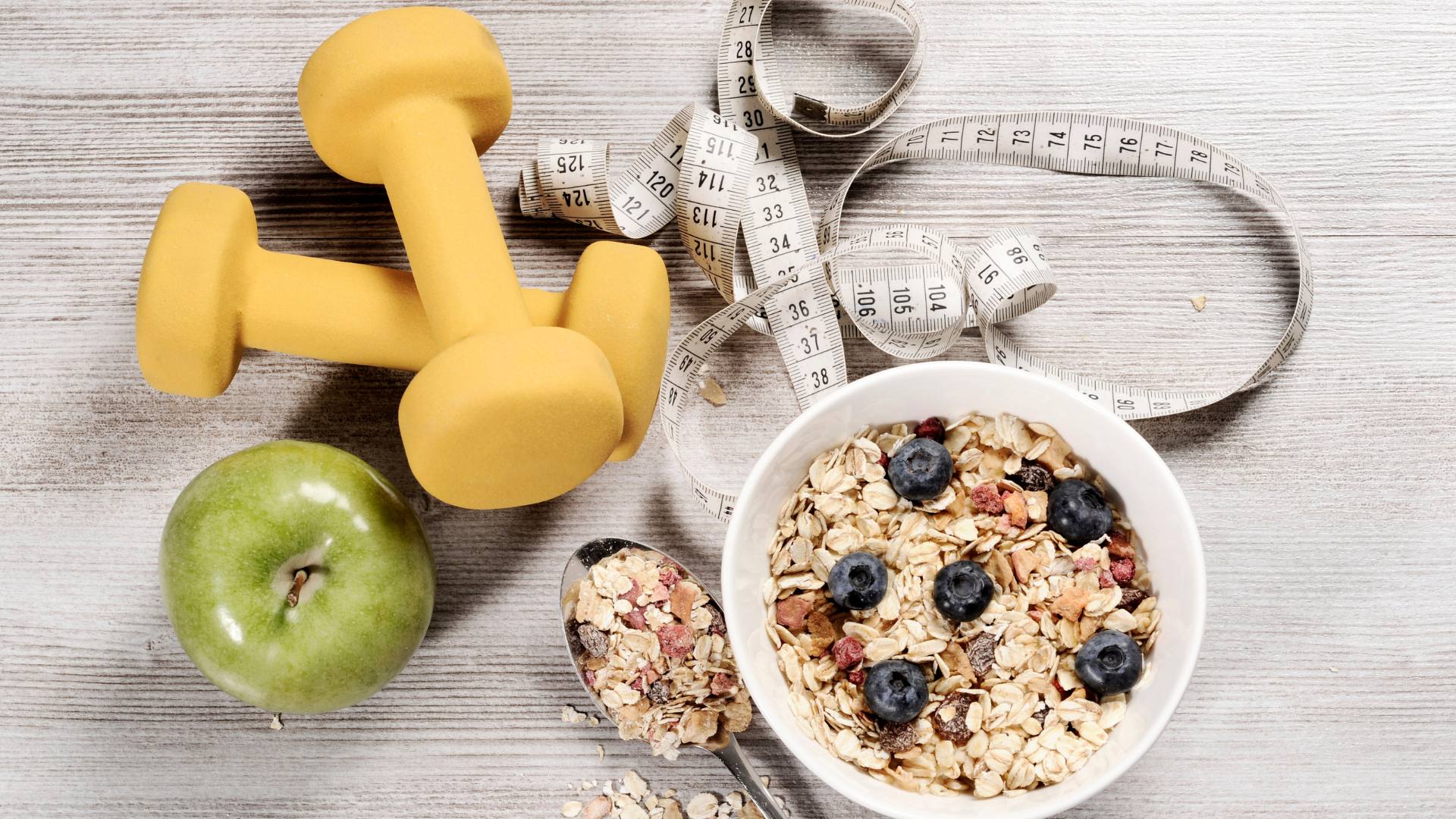 A bowl of cereal is surrounded by an apple, weights, and a measuring tape. This is to demonstrate tips to lose weight.