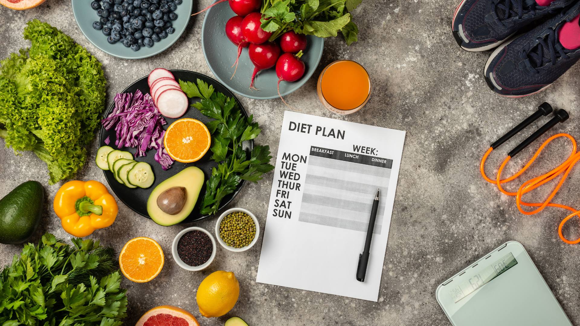 Fruits and vegetables are scattered on a table, accompanied by gym sneakers, a jump rope, a scale, and a notepad with the words "Diet Plan" at the top. These are some tips for losing weight.
