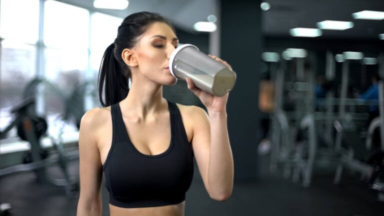 A women wearing a black sports bra wears her hair in a ponytail and sips out of a silver water bottle. She is combatting weight gain.