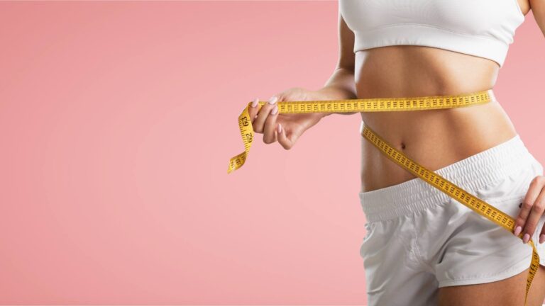 A woman stands wearing white shorts and a white sports bra. Only her torso is visible as she puts a measuring tape around her waist. She is demonstrating her weightloss process.